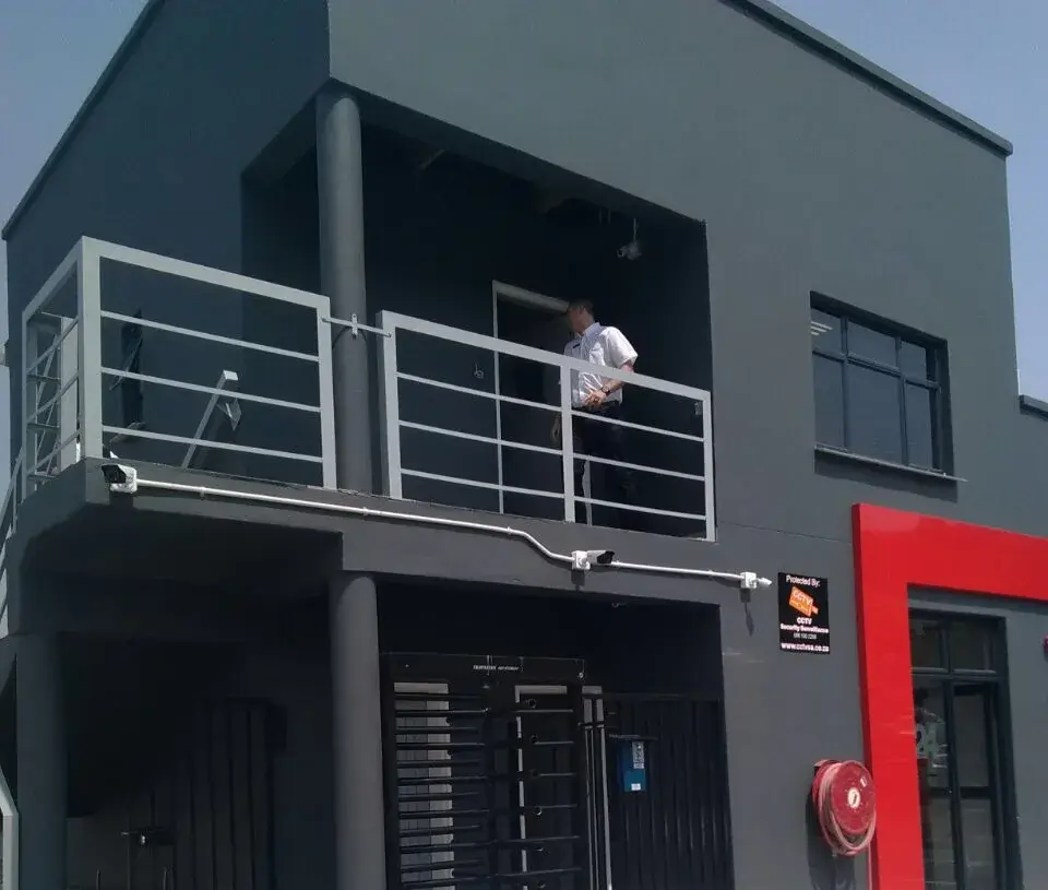 Building entrance protected by CCTV Security Surveillance, with wall-mounted bullet cameras, a full-height access turnstile and a 'Protected by' sign